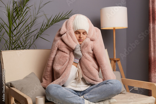 Horizontal shot of cold woman sitting on the sofa at home with crossed legs wearing hat and mittens, wrapped winter coat, trying to get warm in freze apartment without central heating. photo