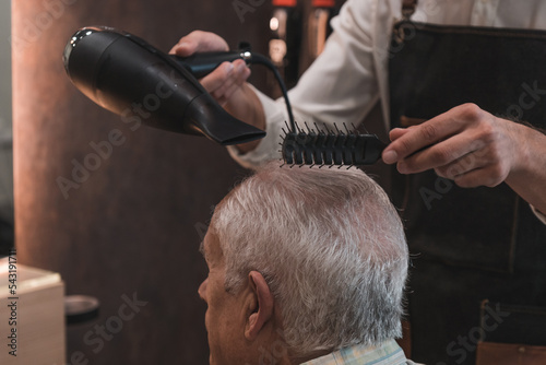 Peluquero irreconocible secando el pelo al cliente senior, con el secador y peine, en la peluquería. photo