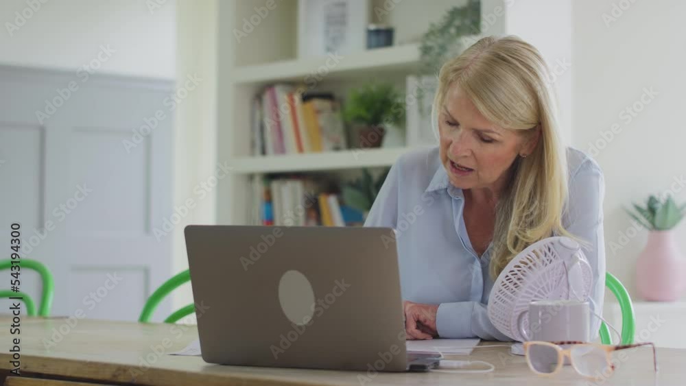 Menopausal mature woman working on laptop with connected fan at home having hot flush and fanning herself with documents - shot in slow motion Menopausal mature woman working on laptop with connected fan at home having hot flush and fanning herself with documents - shot in slow motion