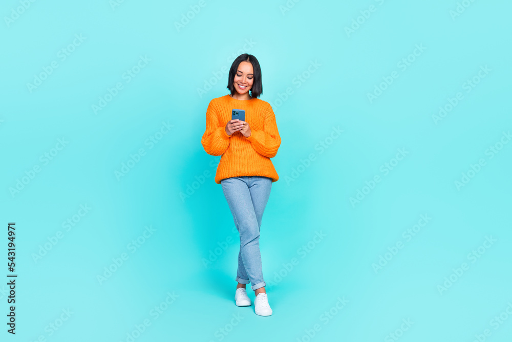 Full length photo of charming adorable girl dressed orange pullover typing modern gadget isolated teal color background Full length photo of charming adorable girl dressed orange pullover typing modern gadget isolated teal color background