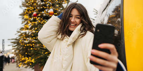 Happy young caucasian girl laughing takes selfie with christmas tree on background. Brunette wears dark blue sweater and jacket. Concept of selfie, modern tachnologies. photo