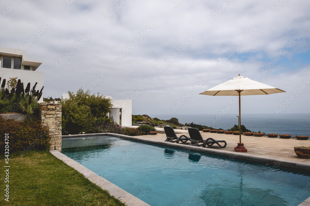 Beautiful garden with grass, big pool and terrace with chairs and parasol and the ocean in a summer evening