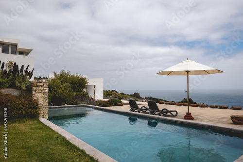 Beautiful garden with grass  big pool and terrace with chairs and parasol and the ocean in a summer evening
