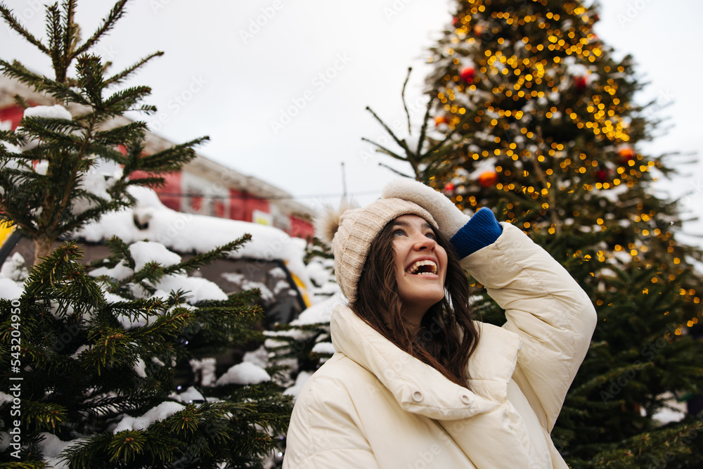 Happy young caucasian woman looking on sky laughing stands outdoor near Christmas tree in winter. Brunette wears hat, jacket and gloves. Positive emotions, weekends concept. Happy young caucasian woman looking on sky laughing stands outdoor near Christmas tree in winter. Brunette wears hat, jacket and gloves. Positive emotions, weekends concept.
