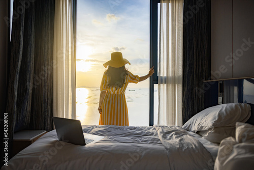 Laptop on white bed sheets in cozy room. Young businesswoman standing at window in confident pose looking at beautiful seascape panorama with rays of sun light in holiday in hotel.Morning in the hotel