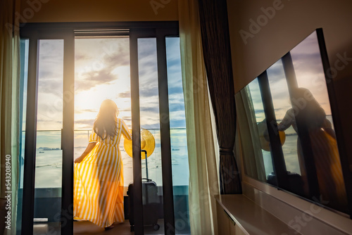 Hotel vacation happy Asian woman tourist in yellow summer dress with her luggage and enjoying ocean view from balcony hotel room and Beautiful sunset above sea or ocean, reflection of sun in the water