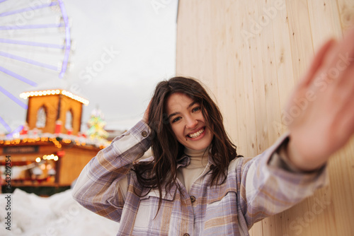 Cheerful young caucasian girl wide smiling at camera streches hand for selfie in winter evening. Brunette wears turtleneck and shirt. Concept of fun, winter holidays. photo