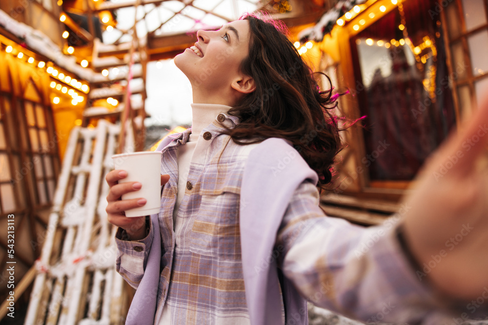 Joyful young caucaian girl looking at sky smiling holds hot coffee in hand spends time outside. Brunette wears golf, shirt and sweater. Concept of positive emotions, lifestyle.