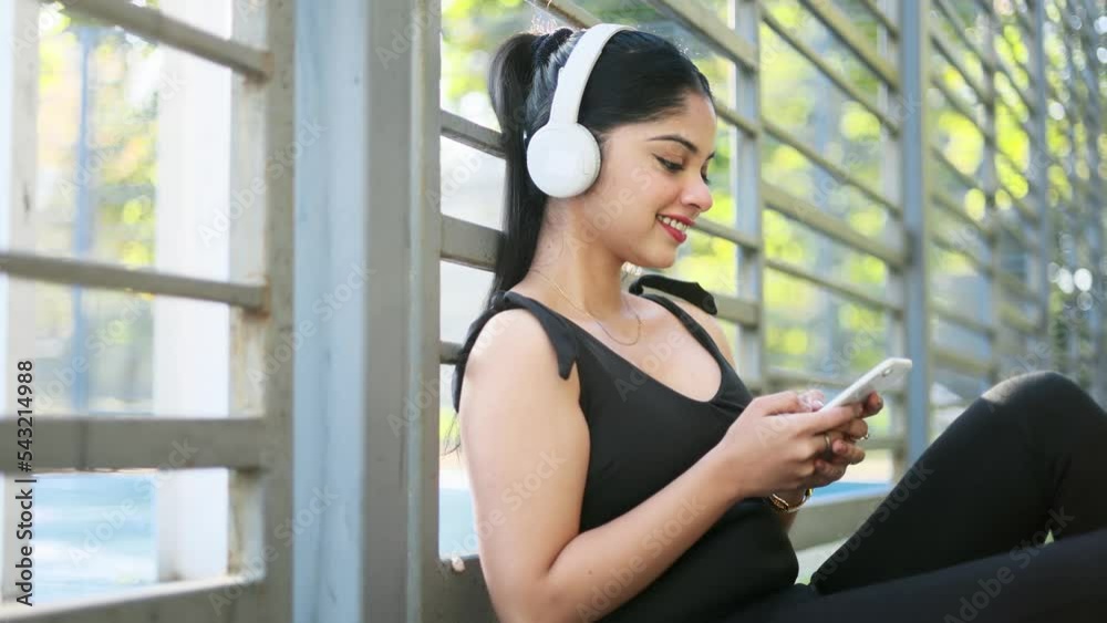 Close up beautiful indian woman hold smartphone, watching social media sitting near a stadium. Smiling young woman with headphones texting on her phone. Close up beautiful indian woman hold smartphone, watching social media sitting near a stadium. Smiling young woman with headphones texting on her phone.