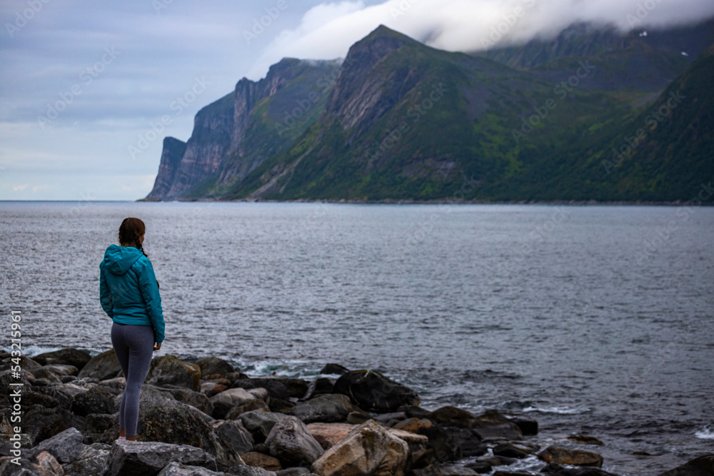 girl in blue jacket stands on a rocky beach admiring the mighty mountains on the island of senja, norway; hiking in the norwegian fjords