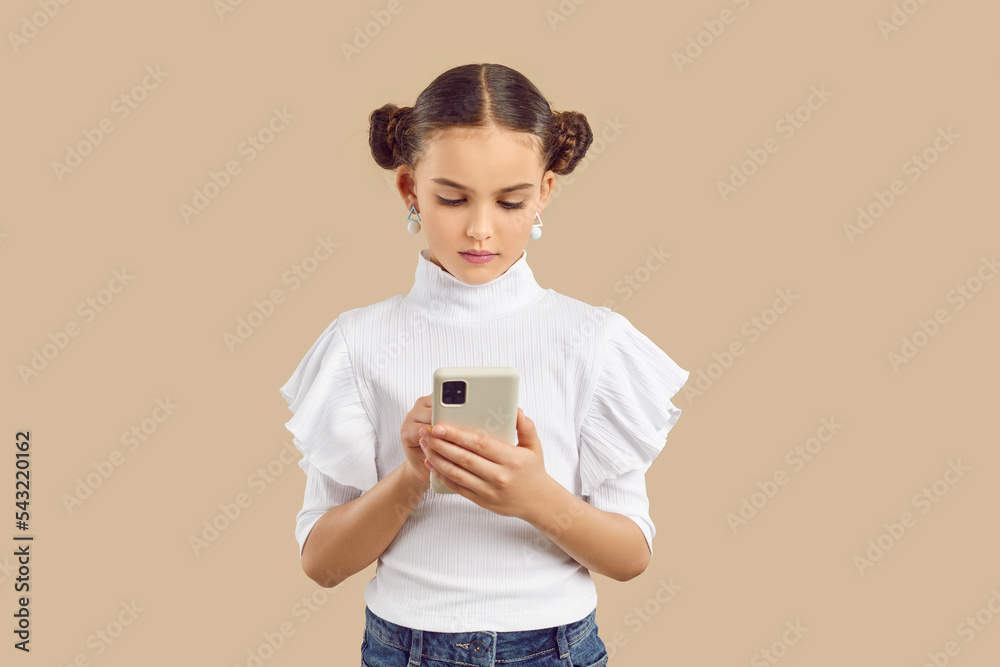 Child using modern smartphone. Little kid texting on mobile phone. Beautiful girl standing on beige studio background, searching information on Internet, reading messages, scrolling social media feed Child using modern smartphone. Little kid texting on mobile phone. Beautiful girl standing on beige studio background, searching information on Internet, reading messages, scrolling social media feed