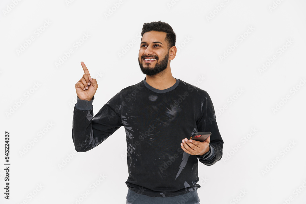 Fototapeta premium Young indian man pointing finger upward and using cellphone