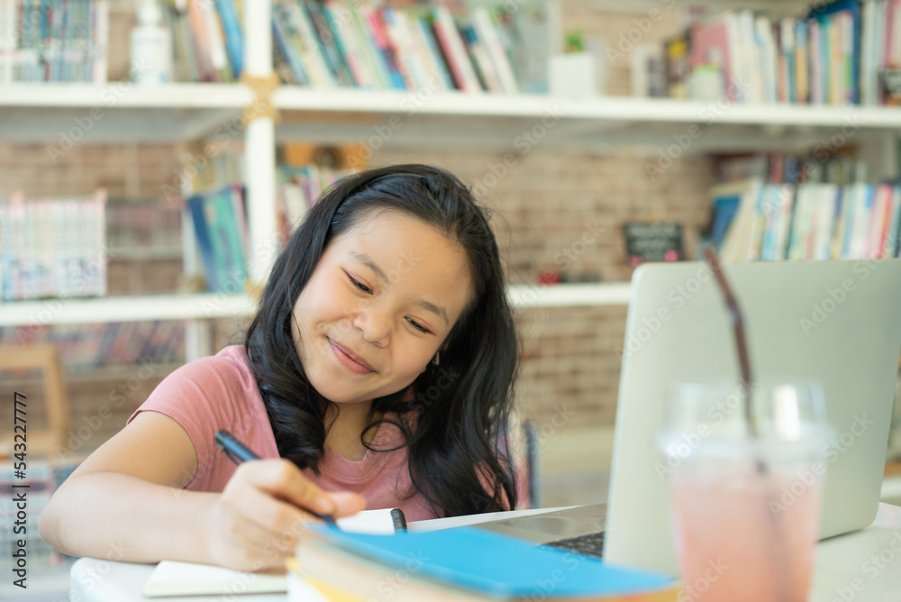 Smiling asian undergraduate teen girl student study in library with laptop books doing online research for coursework, making notes for essay homework assignment, online education e-learning concept.