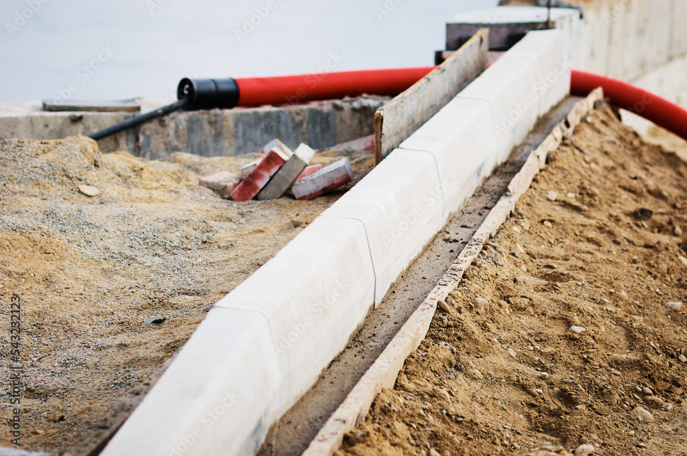 A red corrugated pipe for laying an electrical cable sticks out of the ground at a construction site. Curb stones are installed in a row using formwork and liquid concrete. Embankment construction A red corrugated pipe for laying an electrical cable sticks out of the ground at a construction site. Curb stones are installed in a row using formwork and liquid concrete. Embankment construction