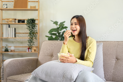 Young asian woman putting a bowl of popcorn on pillow and eating popcorn while sitting on the big comfortable