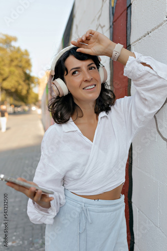 Obraz na płótnie European cute lovely woman with dark wavy short hair wearing white blouse and blue skirt in headphone is holding smartphone, raised up hands and looking aside on the street w salonie