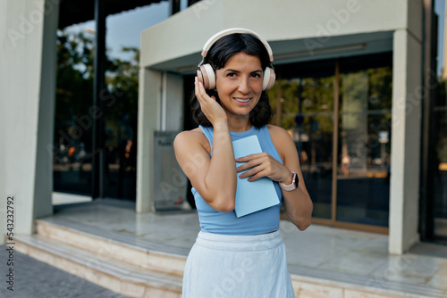 Pretty attractive woman with short dark wavy hair wearing summer blue t-shirt and white skirt listening music in headphones and looking at camera 