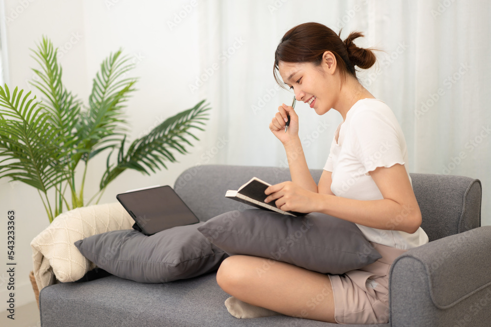 Young asian woman writing new idea of project in notebook and working on tablet while wearing casual Young asian woman writing new idea of project in notebook and working on tablet while wearing casual
