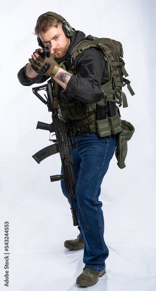 Fototapeta premium an airsoft player in full gear pointed the muzzle of a pistol at the target. a man in an outfit, in headphones, a bulletproof vest, with a backpack and a belt with additional shells. white background.