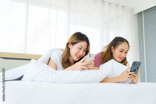 Two young Asian women friends using smart phones while lying down with a pillow and blanket on the bed together. LGBTQIA girl-friendly friendship sharing happy times together in the bedroom.