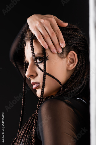 portrait of stylish woman with makeup and piercing touching braids near white wall on black background.