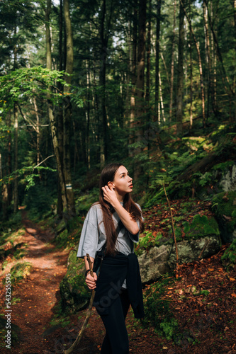 Beautiful woman in casual clothes stands in the middle of the forest in the mountains and rests on a hike in the sun. © bodnarphoto