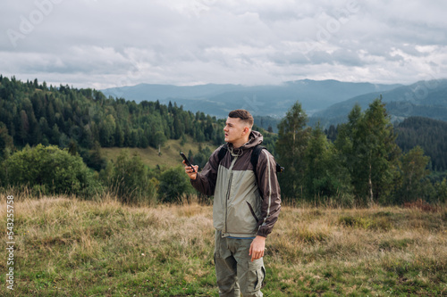 A handsome young male tourist in casual clothes stands in the mountains with a smartphone in his hands and looks at the route where to go next.