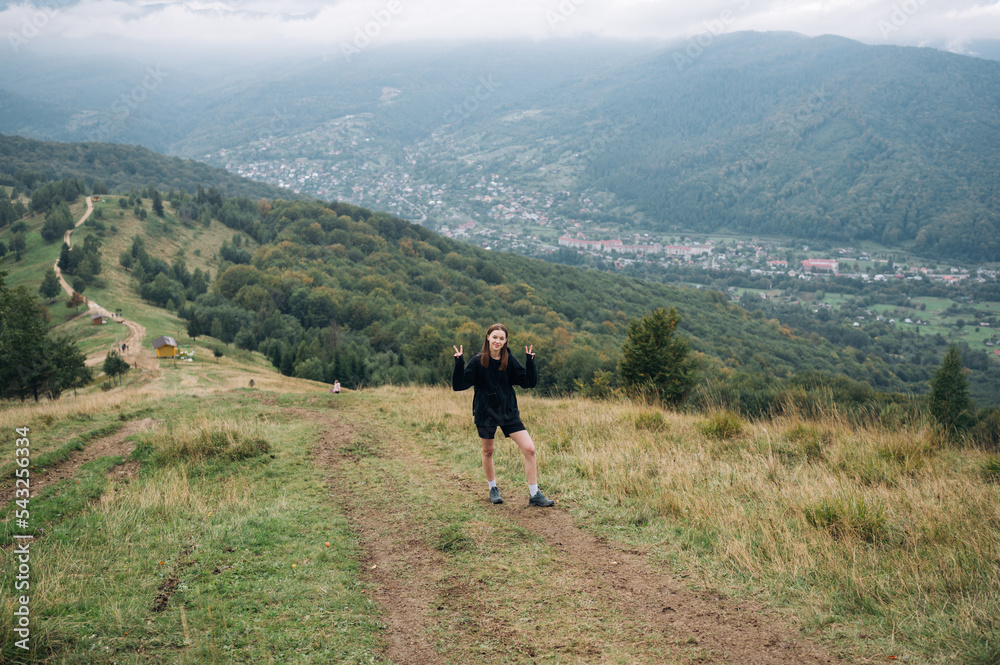 Funny tourist girl in dark clothes stands on the path under the chaas climbing the mountain against the background of beautiful views and poses for the camera, shows a peace gesture.