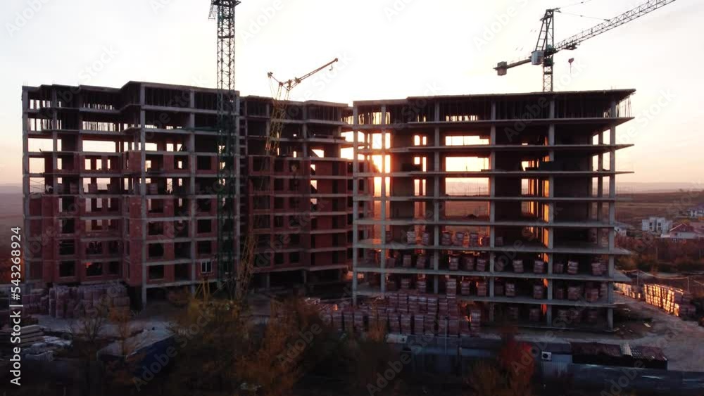Aerial Flight Over a New Constructions Development Site with High Tower Cranes Building Real Estate. Heavy Machinery and Construction Workers are Employed. Blue Sea and City Houses in the Background.