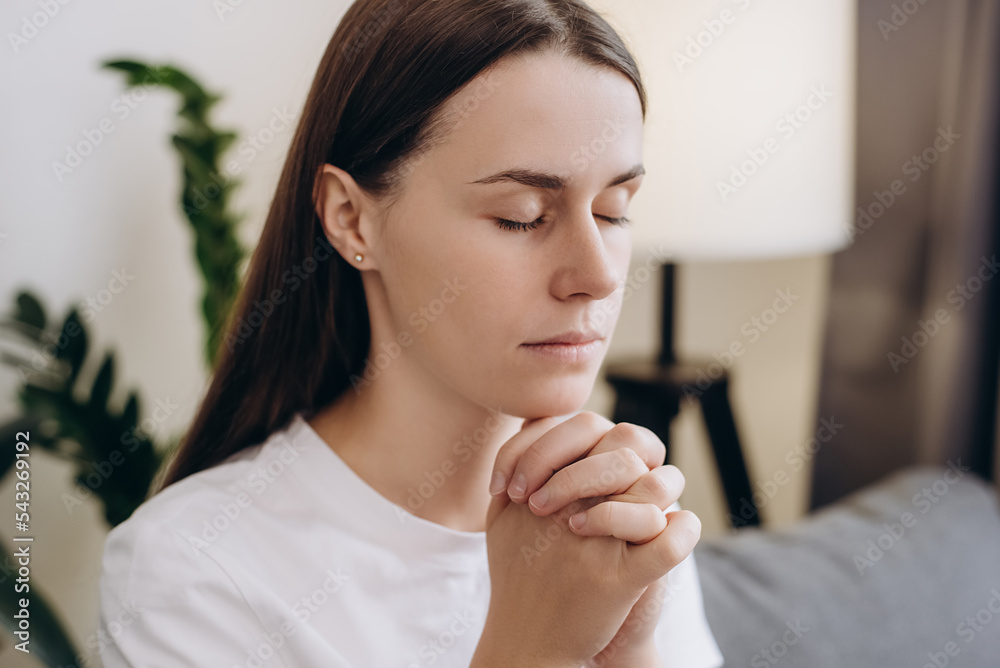 Cute girl praying sincerely with folded arms and making wish, asking with hopeful imploring expression, begging apology. Calm young woman sitting on couch at home living room. Please, God help. Cute girl praying sincerely with folded arms and making wish, asking with hopeful imploring expression, begging apology. Calm young woman sitting on couch at home living room. Please, God help.