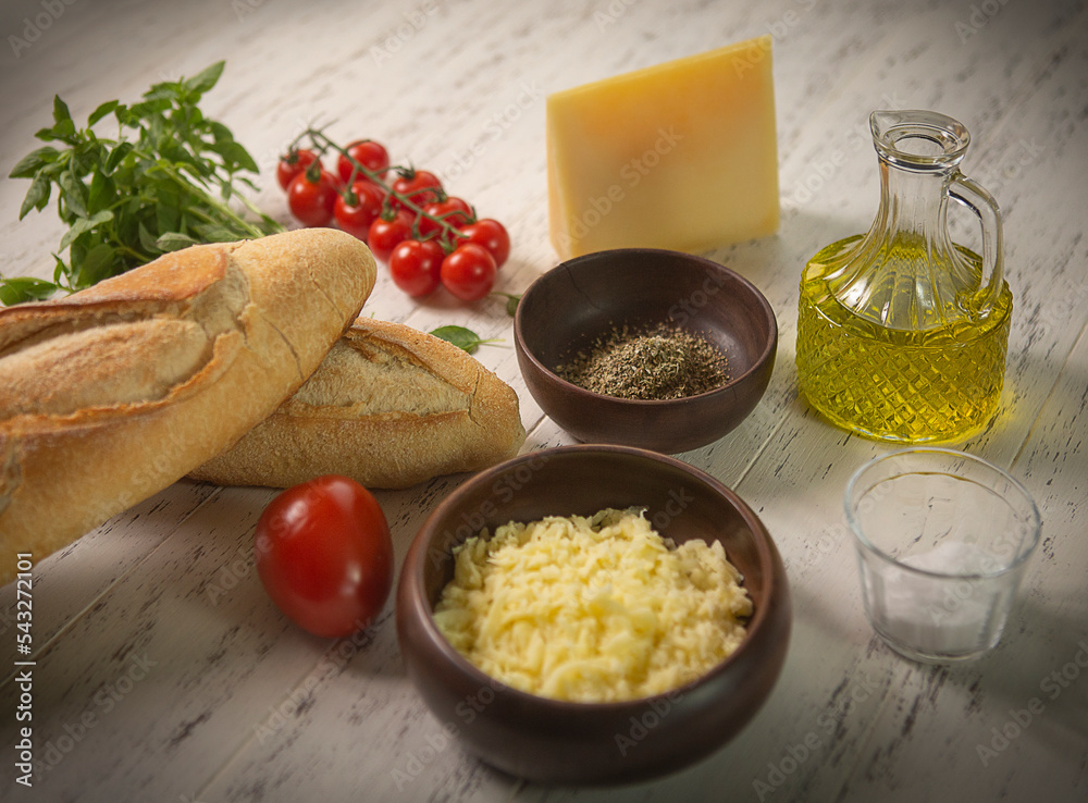 Ingredientes para brusqueta, pão italiano, tomates, tomates cereja, queijo mussarela, ervas finas, queijo parmesão, azeite de oliva e manjericão, em fundo de mesa branco manchado. Foco seletivo.
