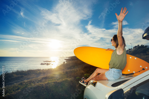 Woman with surfboard on car hood look over sunset lift hand up
