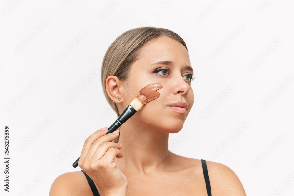 Young caucasian blonde woman applying the tonal foundation to the face with a brush isolated on a white background. A thick smear of a clay mask on the girl's cheek Young caucasian blonde woman applying the tonal foundation to the face with a brush isolated on a white background. A thick smear of a clay mask on the girl's cheek