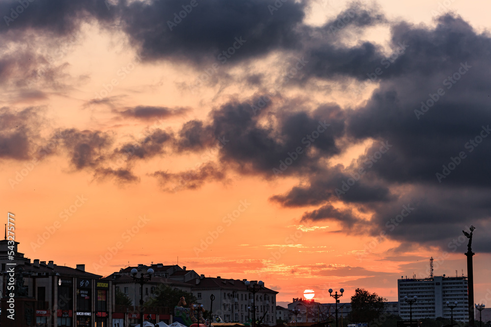 Orange sunset with sun disk, clouds, roofs of buildings.