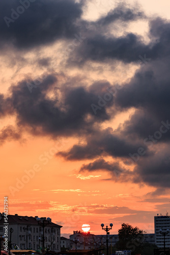 Orange sunset with sun disk, clouds, roofs of buildings. Vertical orientation.