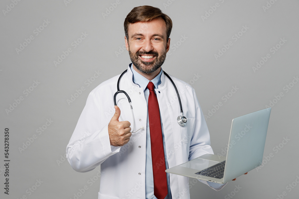 Male doctor smiling happy IT man wearing white medical gown suit stethoscope work in hospital hold use laptop pc computer show thumb up isolated on plain grey background. Healthcare medicine concept. Male doctor smiling happy IT man wearing white medical gown suit stethoscope work in hospital hold use laptop pc computer show thumb up isolated on plain grey background. Healthcare medicine concept.