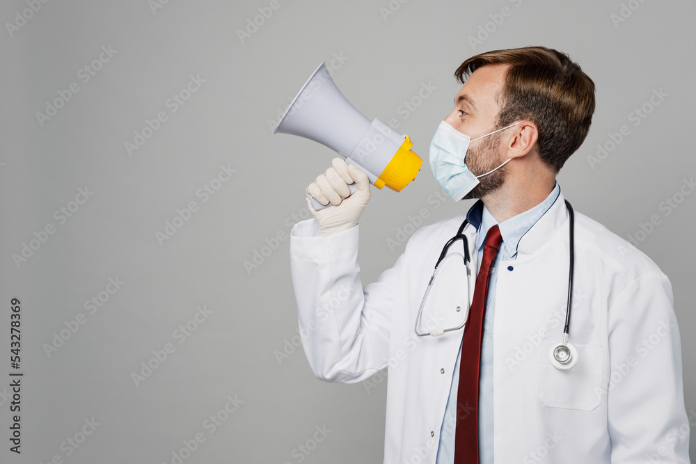 Male doctor man wearing white medical gown suit sterile mask work in hospital scream aside in megaphone bullhorn isolated on plain grey color background studio portrait. Healthcare medicine concept. Male doctor man wearing white medical gown suit sterile mask work in hospital scream aside in megaphone bullhorn isolated on plain grey color background studio portrait. Healthcare medicine concept.