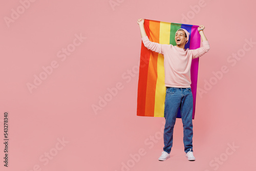 Full body young gay man wear sweatshirt hat hold striped flag behind himself look aside on workspace isolated on plain pastel light pink color background studio portrait Lifestyle lgbtq pride concept photo