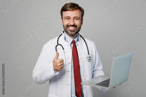 Male doctor smiling happy IT man wearing white medical gown suit stethoscope work in hospital hold use laptop pc computer show thumb up isolated on plain grey background. Healthcare medicine concept.