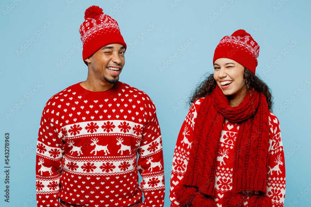 Merry young positive fun couple friends two man woman wear red Christmas sweater Santa hat posing look camera wink isolated on plain pastel light blue background Happy New Year 2023 holiday concept