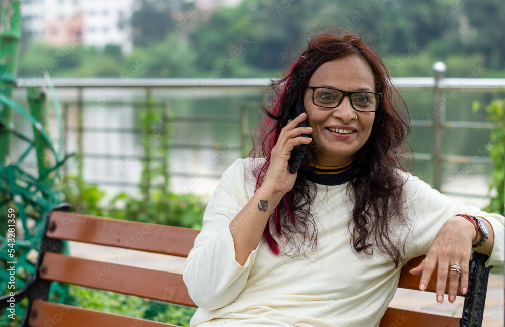 Smiling Woman with long brown hair Speaking on mobile phone relaxing outdoors. Closeup. She is wearing White T-shirt and specs. Front view. Head and Shoulder Shot. Smiling Woman with long brown hair Speaking on mobile phone relaxing outdoors. Closeup. She is wearing White T-shirt and specs. Front view. Head and Shoulder Shot.