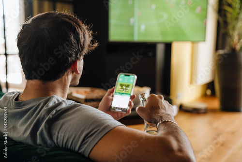 Back view of man watching football match and making bets at bookmaker's website in front of TV screen photo