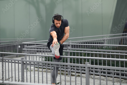 Young man in sports clothes and headphones on his head stretching legs before jogging