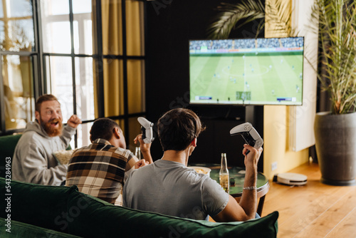 Back view of male friends playing football video game with gamepads at home photo