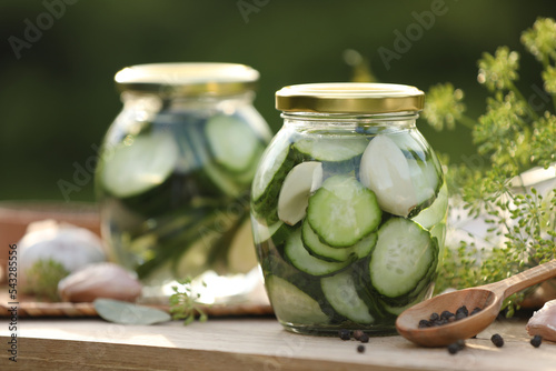 Jars of delicious pickled cucumbers and ingredients on wooden table against blurred background, closeup. Space for text