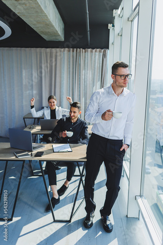 A successful manager man in a white shirt stands by the window against the background of his colleagues in a modern office. Working atmosphere in the office.