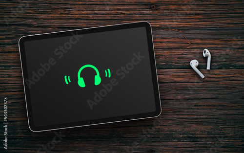 Green headphones on dark screen of tablet and wireless earphones on dark wooden surface