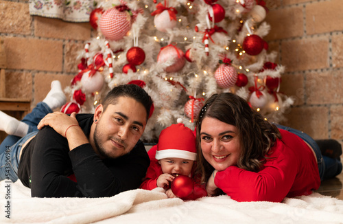 happy mulit ethnic family hispanic caucasian young smiling couple with a baby in front of white cristmas tree, mom with red christmas sweater and dad wearing black cristmas sweater lying on the floor photo