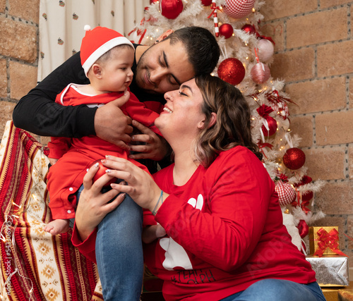 happy mulit ethnic family hispanic caucasian young smiling couple with a baby in front of a white cristmas tree, mom with red christmas sweater and dad wearing black cristmas sweater on a couch