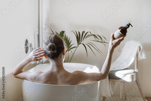 Young woman taking bath, holding bottle with shower gel in hand photo